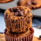 Two chocolate chip muffins stacked on each other, with chocolate chips on top. A muffin tin and some scattered chocolate chips are in the background.