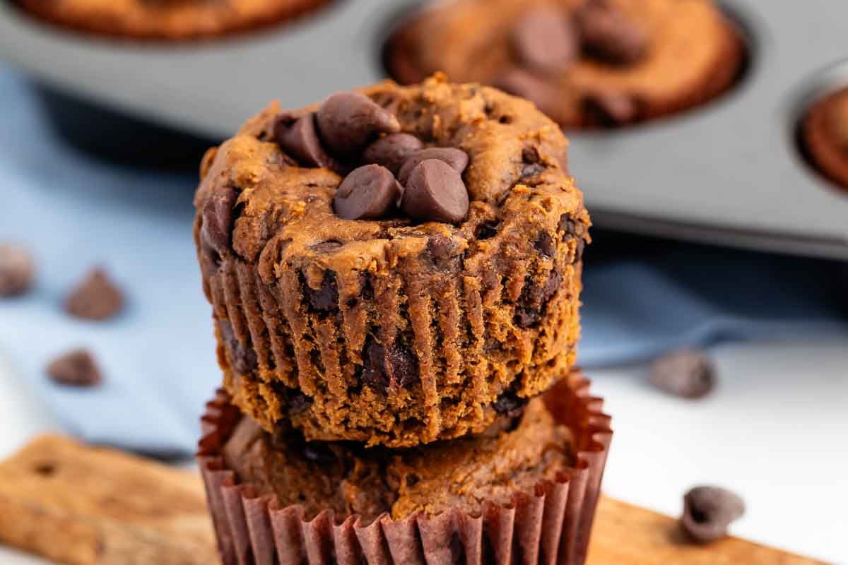 Two chocolate chip muffins stacked on each other, with chocolate chips on top. A muffin tin and some scattered chocolate chips are in the background.