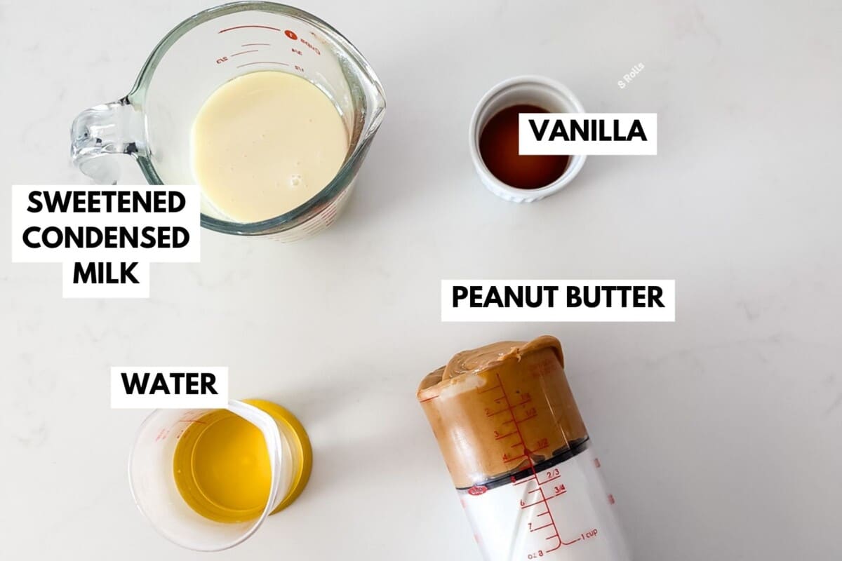 A glass measuring cup with condensed milk, a small cup of vanilla, a cup of water, and a measuring cup of peanut butter are labeled on a white surface.