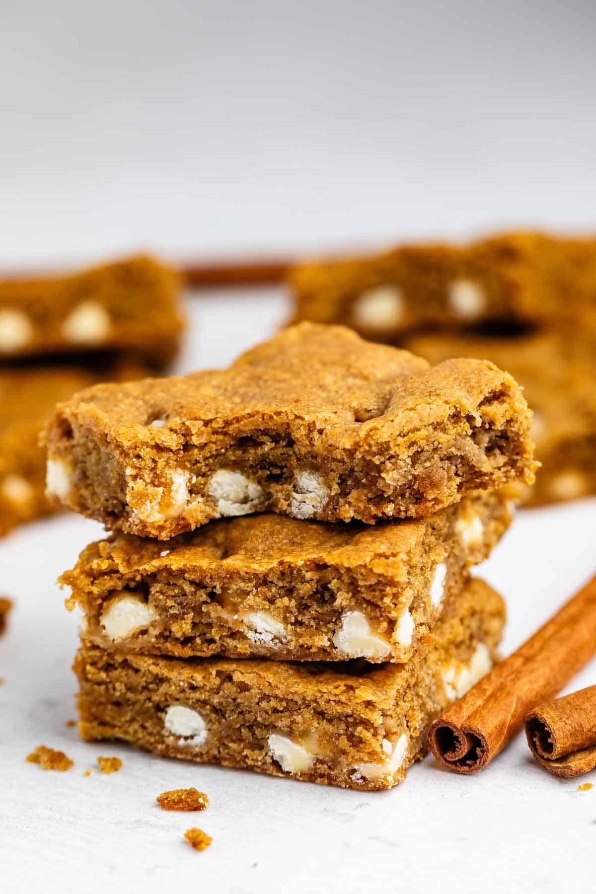 Three stacked pumpkin blondies with white chocolate chips, surrounded by more blondies and two cinnamon sticks on a white surface.