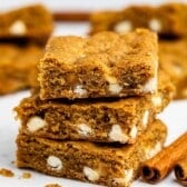 Three blondie bars with white chocolate chips are stacked on a white surface, surrounded by more bars and two cinnamon sticks in the background. The cookies look chewy and golden brown.