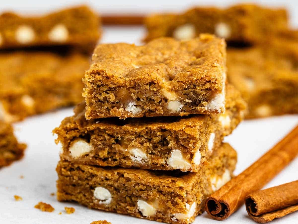 Three blondie bars with white chocolate chips are stacked on a white surface, surrounded by more bars and two cinnamon sticks in the background. The cookies look chewy and golden brown.