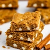 Three stacked blondies with white chocolate chips, surrounded by more blondies and cinnamon sticks on a white surface.