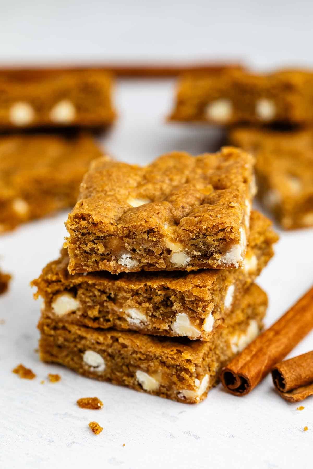 Three stacked blondies with white chocolate chips, surrounded by more blondies and cinnamon sticks on a white surface.