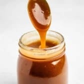 A glass jar filled with thick caramel sauce, with a spoon dipped in and caramel dripping back into the jar.