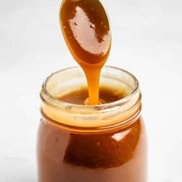 A glass jar filled with thick caramel sauce, with a spoon dipped in and caramel dripping back into the jar.