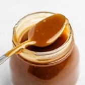 A spoon with caramel sauce rests on the rim of an open glass jar filled with caramel sauce, set against a white background.