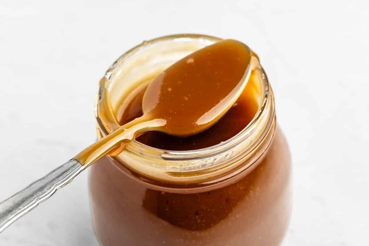 A spoon with caramel sauce rests on the rim of an open glass jar filled with caramel sauce, set against a white background.