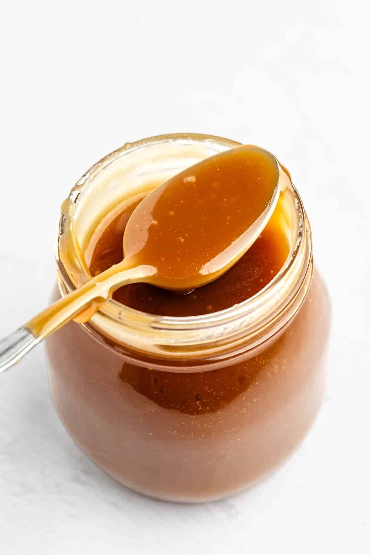 A spoon covered in caramel sauce rests on top of an open glass jar filled with more caramel sauce, set on a white surface.