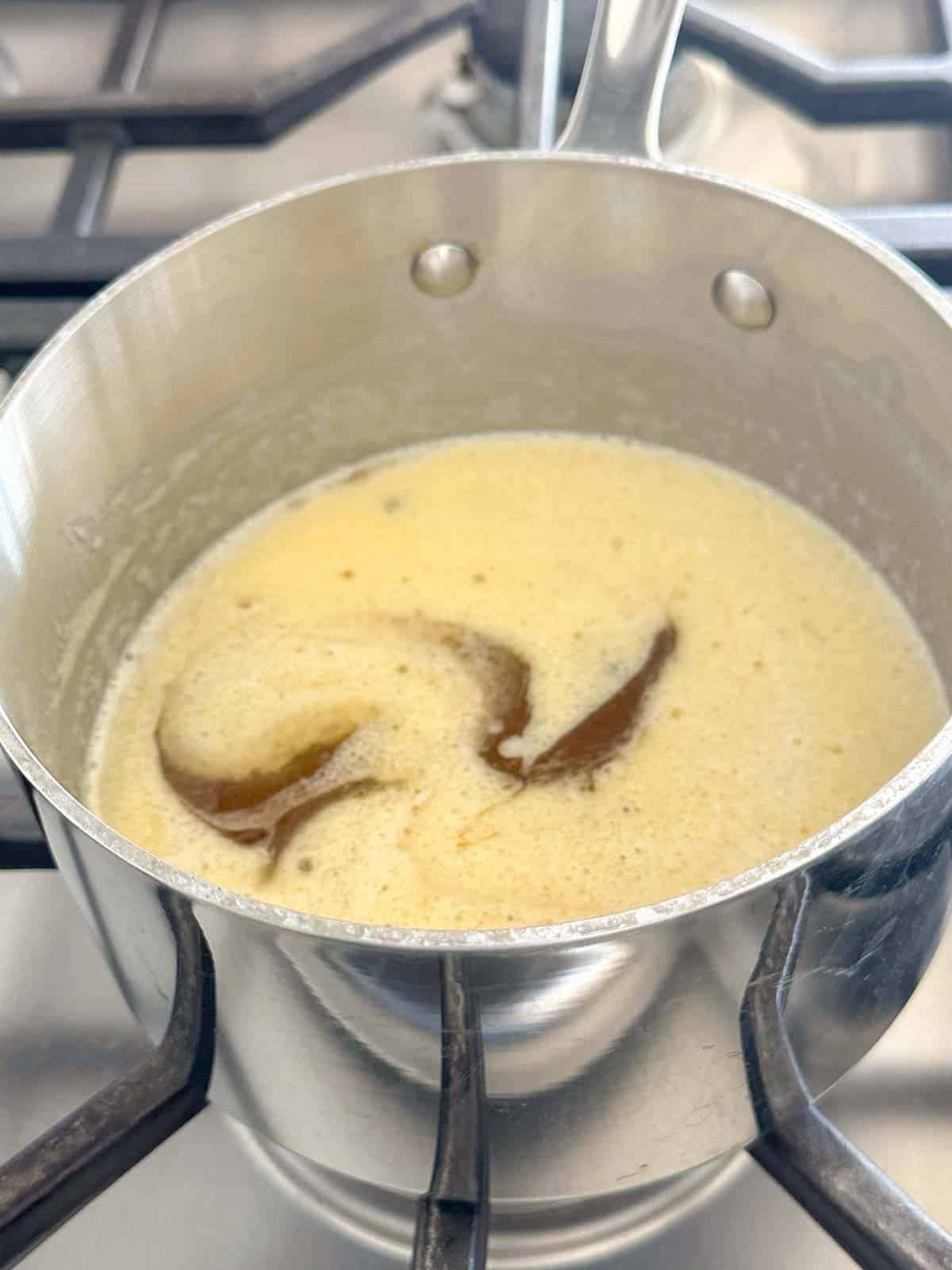 A stainless steel saucepan on a stove contains melting butter, with visible swirls of brown as it begins to brown.