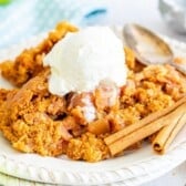 A plate of apple crumble topped with a scoop of vanilla ice cream, with two cinnamon sticks on the side and a spoon resting nearby.