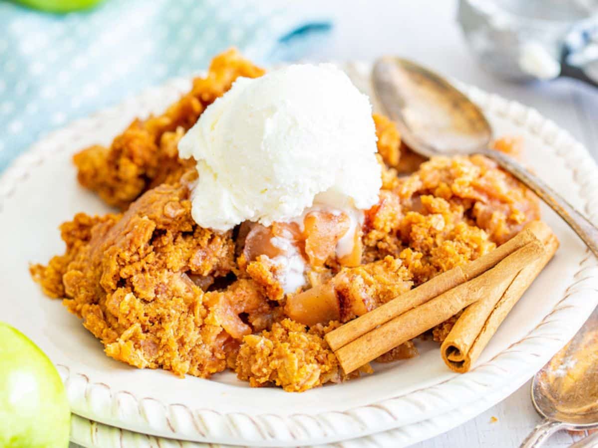 A plate of apple crumble topped with a scoop of vanilla ice cream, with two cinnamon sticks on the side and a spoon resting nearby.