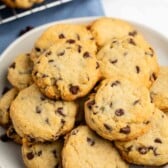 A plate of chocolate chip cookies sits on a white surface, with more cookies on a cooling rack in the background. The cookies are golden-brown and studded with chocolate chips.