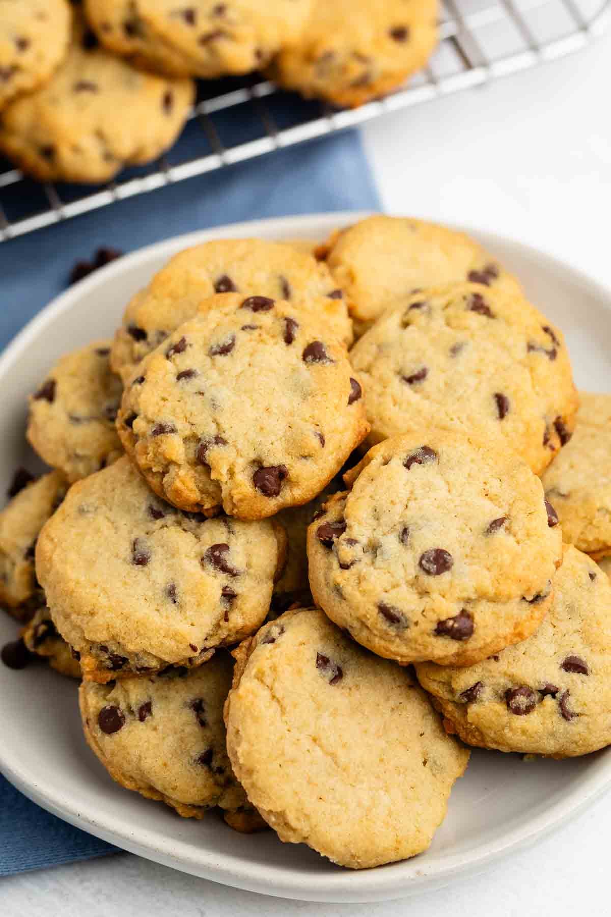 A plate of chocolate chip cookies sits on a white surface, with more cookies on a cooling rack in the background. The cookies are golden-brown and studded with chocolate chips.