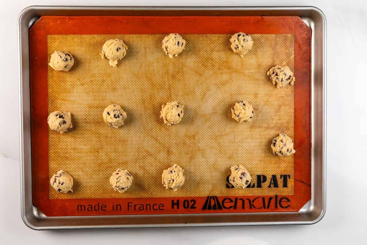 A baking sheet lined with a silicone mat holds 12 evenly spaced scoops of chocolate chip cookie dough, ready to be baked.