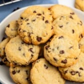 A plate filled with homemade chocolate chip cookies, golden brown and studded with chocolate chips, sits on a table with a few cookies in the background.