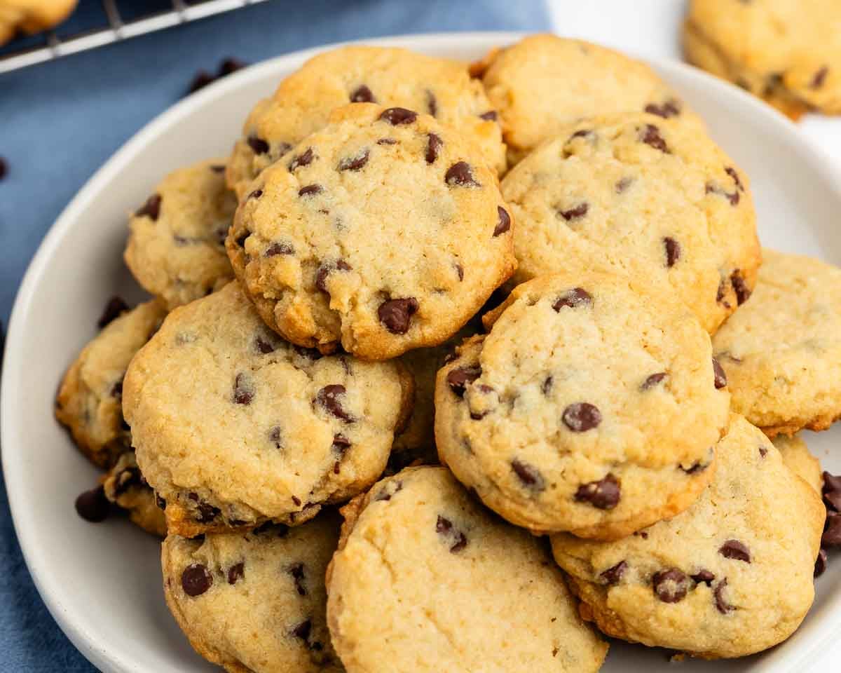 A plate filled with homemade chocolate chip cookies, golden brown and studded with chocolate chips, sits on a table with a few cookies in the background.