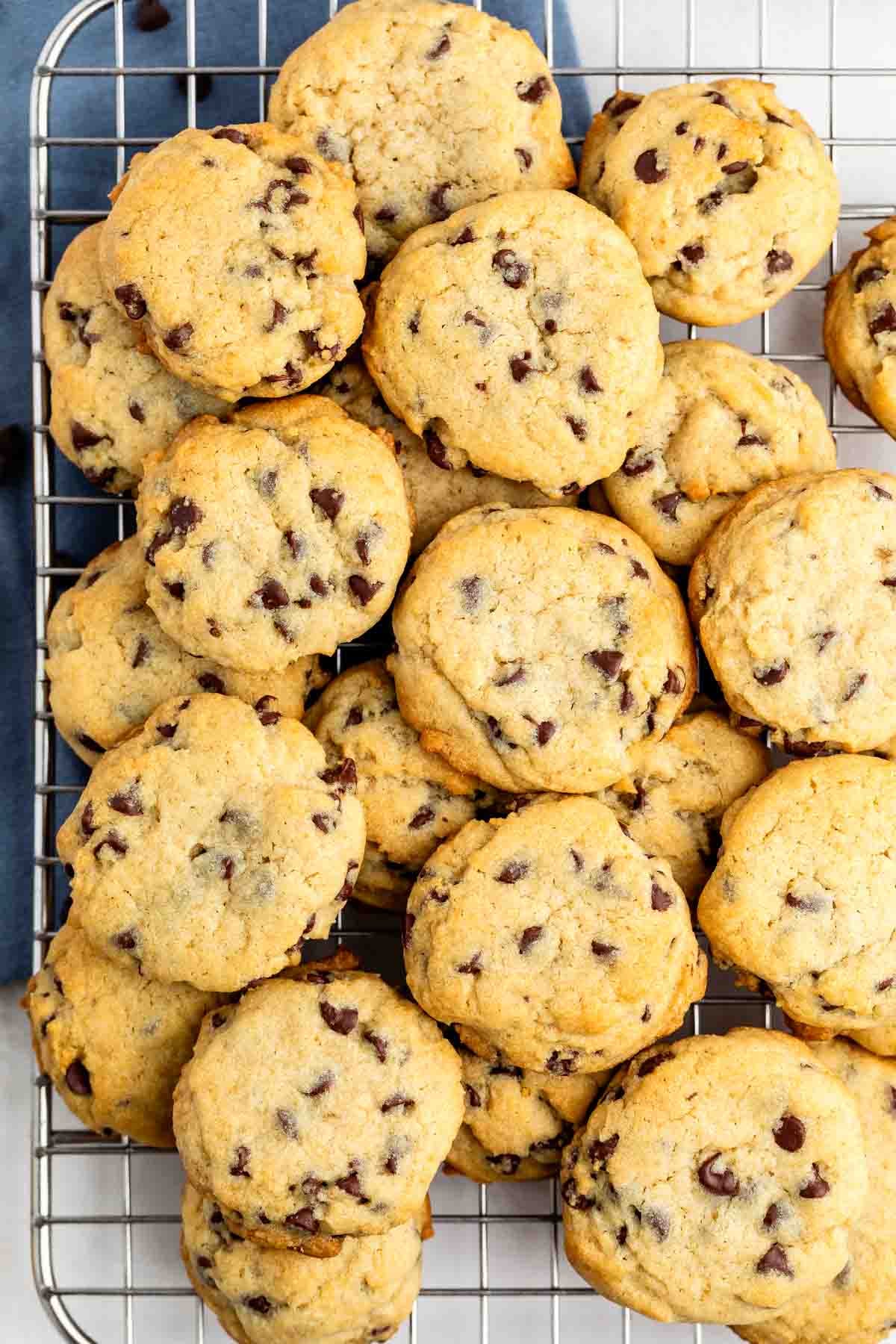 A pile of chocolate chip cookies rests on a wire cooling rack, with golden edges and visible chocolate chips throughout each cookie.