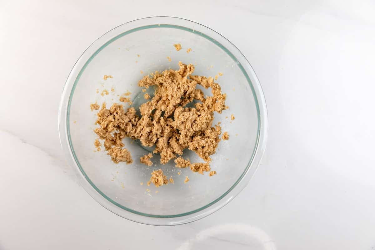 A glass bowl containing a small amount of light brown, crumbly dough sits on a white surface.