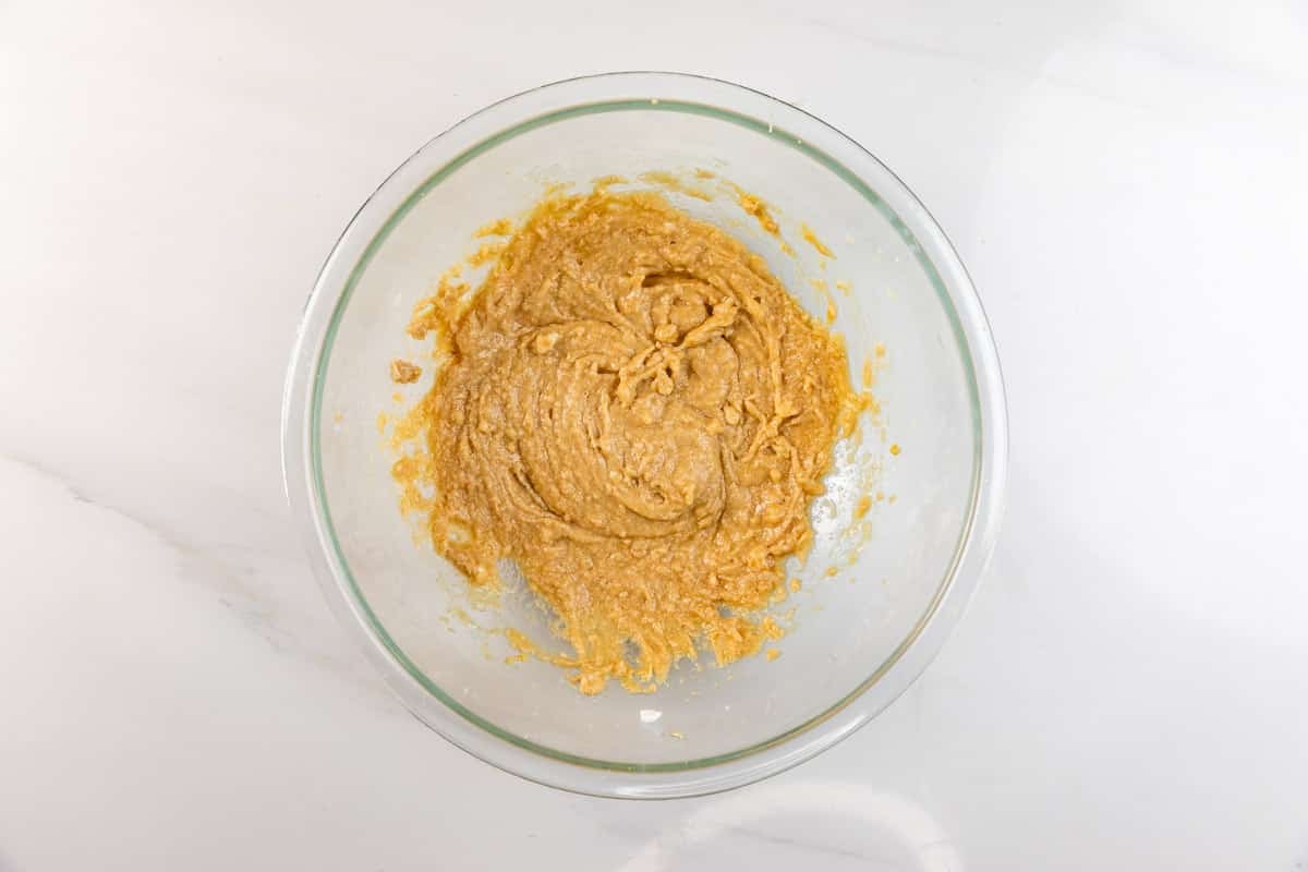 A glass bowl on a white countertop contains thick, light brown batter or dough, partially mixed with some streaks visible along the sides of the bowl.