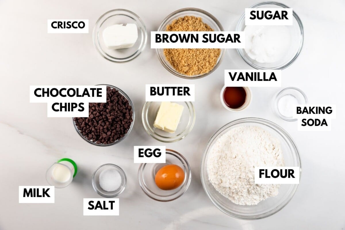 Ingredients for baking are arranged on a white surface in glass bowls: butter, Crisco, brown sugar, sugar, vanilla, flour, baking soda, chocolate chips, salt, milk, and an egg. Each item is labeled.