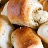A close-up of golden brown, freshly baked garlic rolls topped with herbs and specks of seasoning, arranged in a pile.