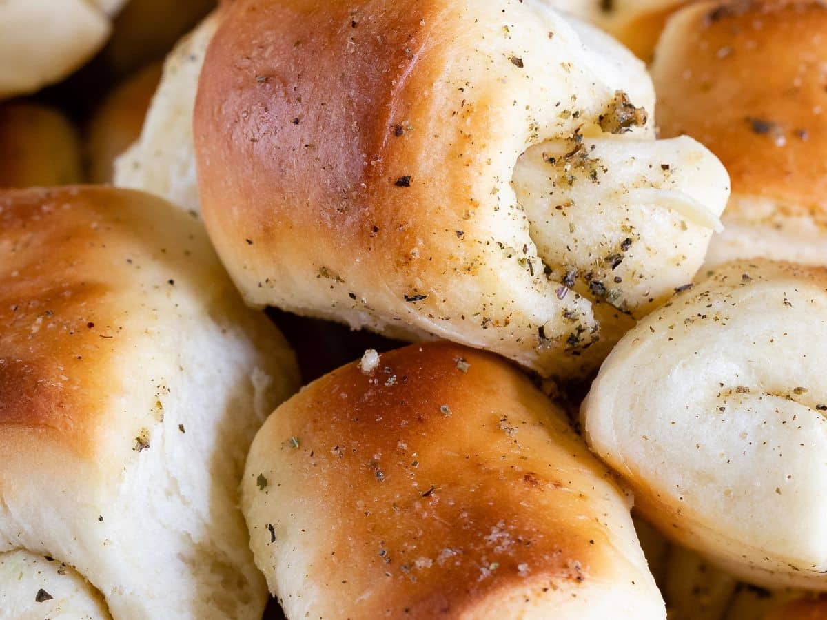 A close-up of golden brown, freshly baked garlic rolls topped with herbs and specks of seasoning, arranged in a pile.