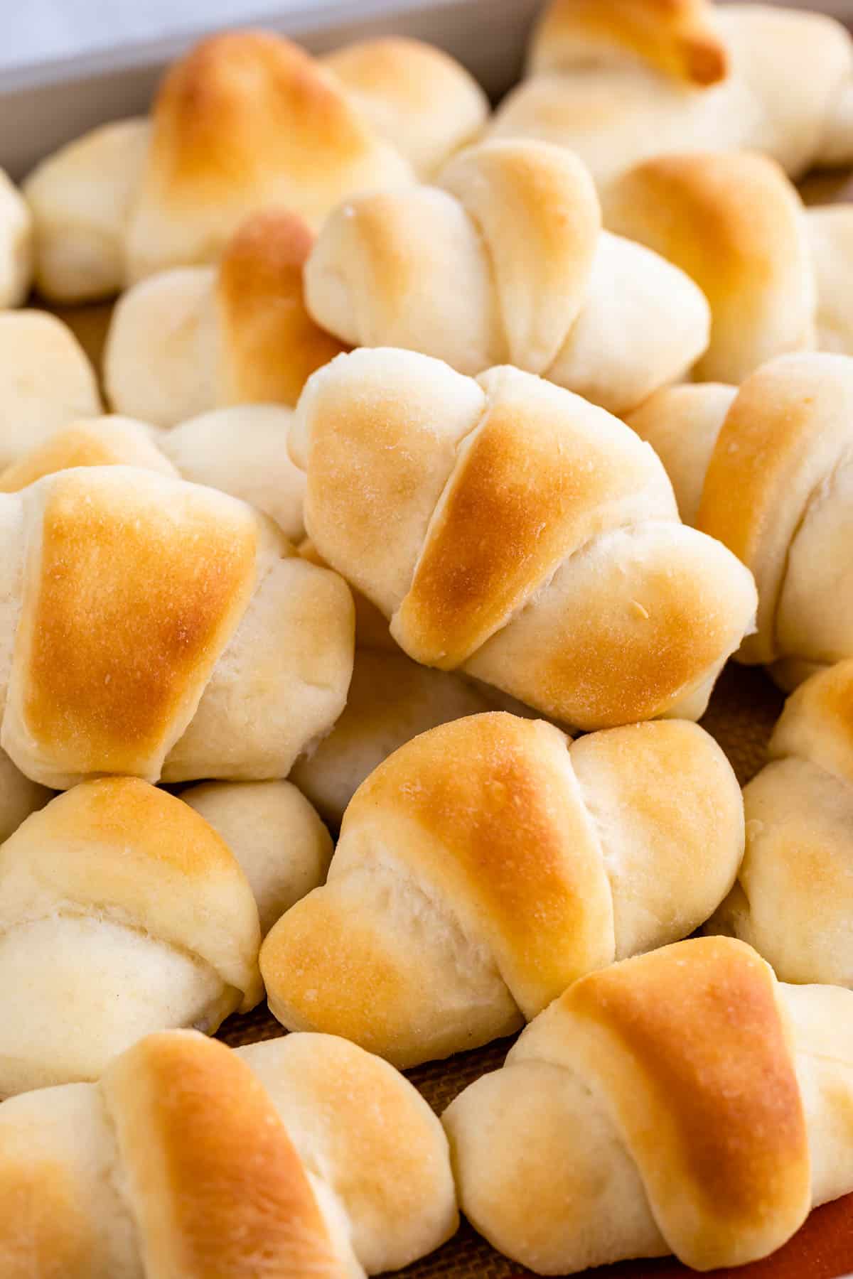 A pile of freshly baked golden brown crescent rolls stacked closely together, showing their soft texture and slightly crisp tops.