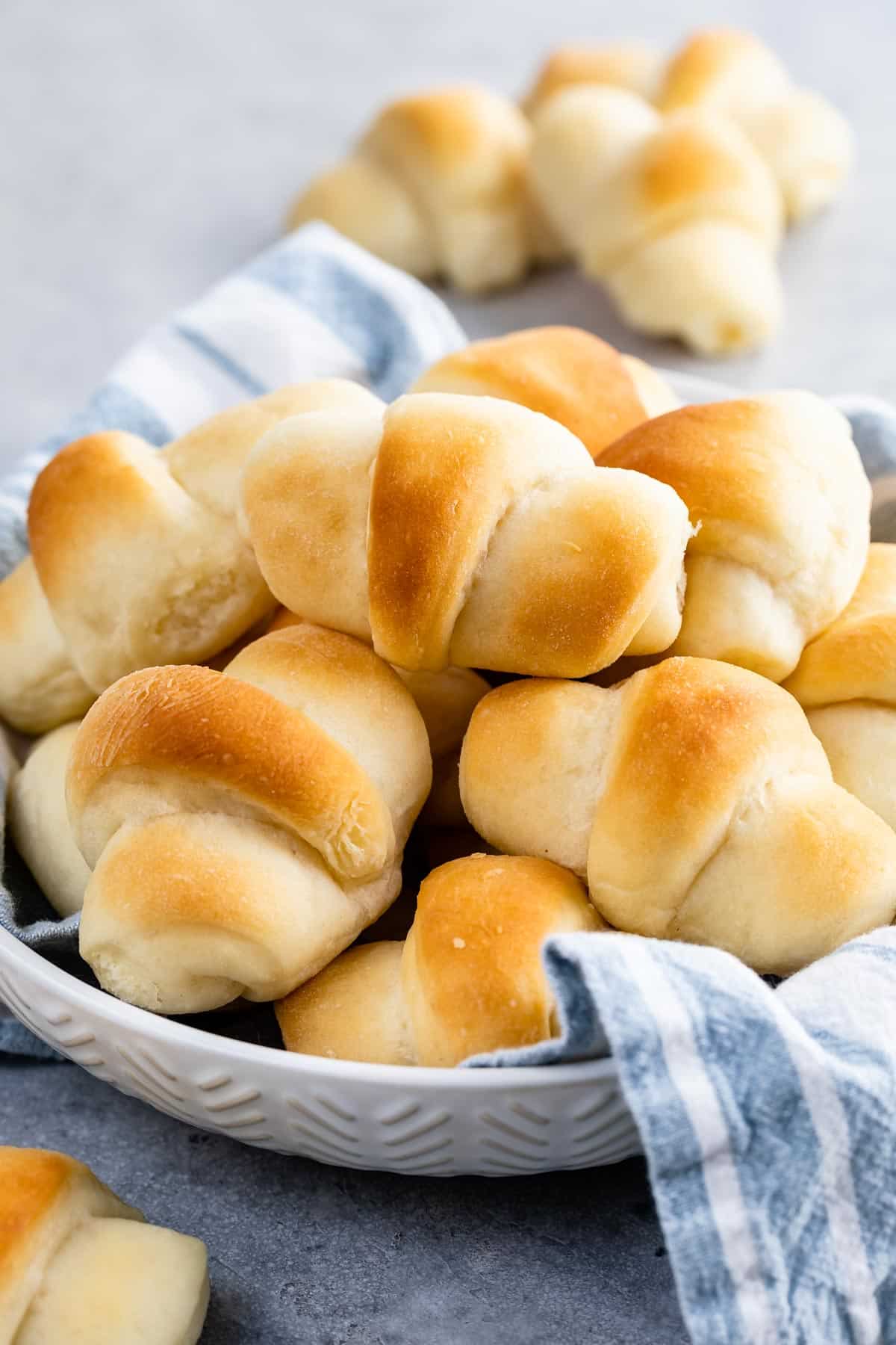 A white dish filled with golden brown dinner rolls, partially wrapped in a blue and white striped cloth, with more rolls in the background on a gray surface.