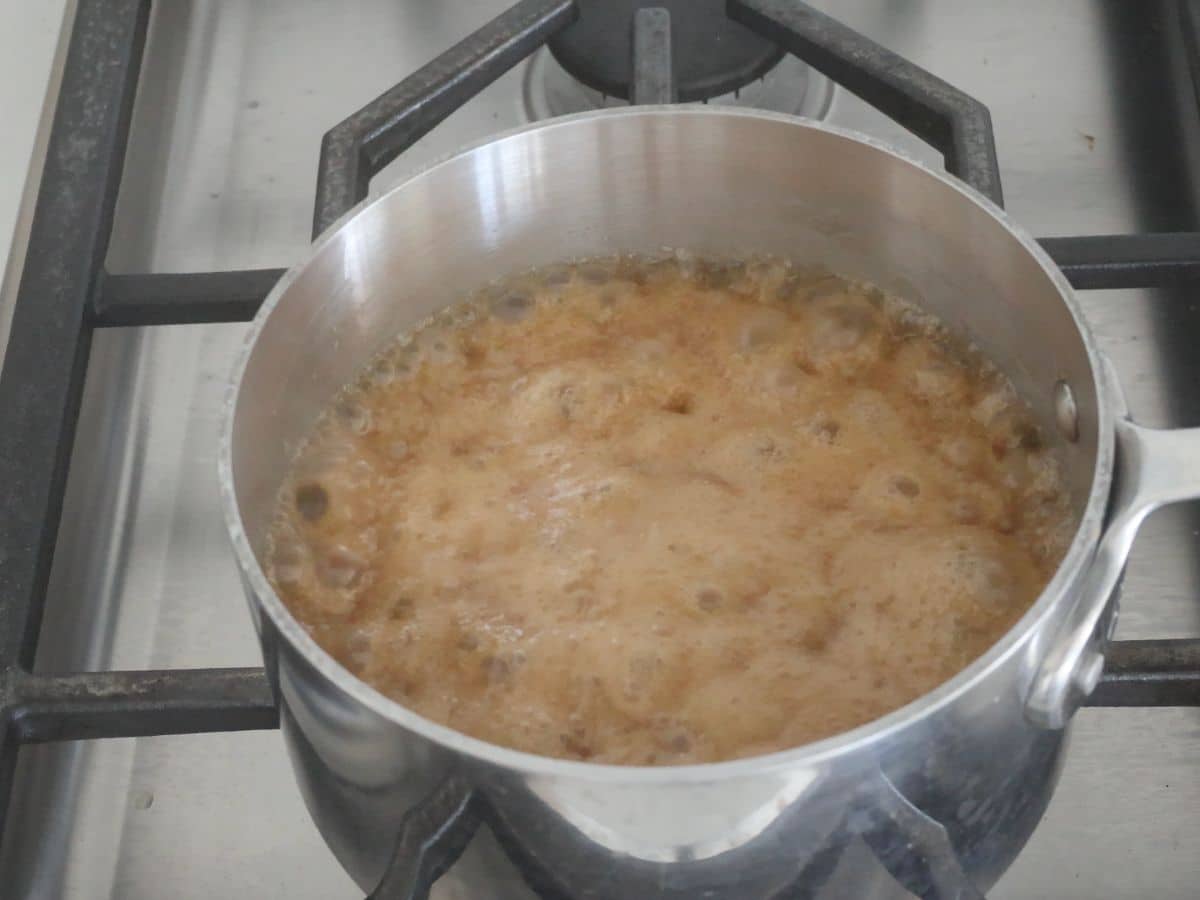 A stainless steel pot filled with bubbling brown liquid sits on a gas stove, likely caramelizing sugar or boiling syrup.