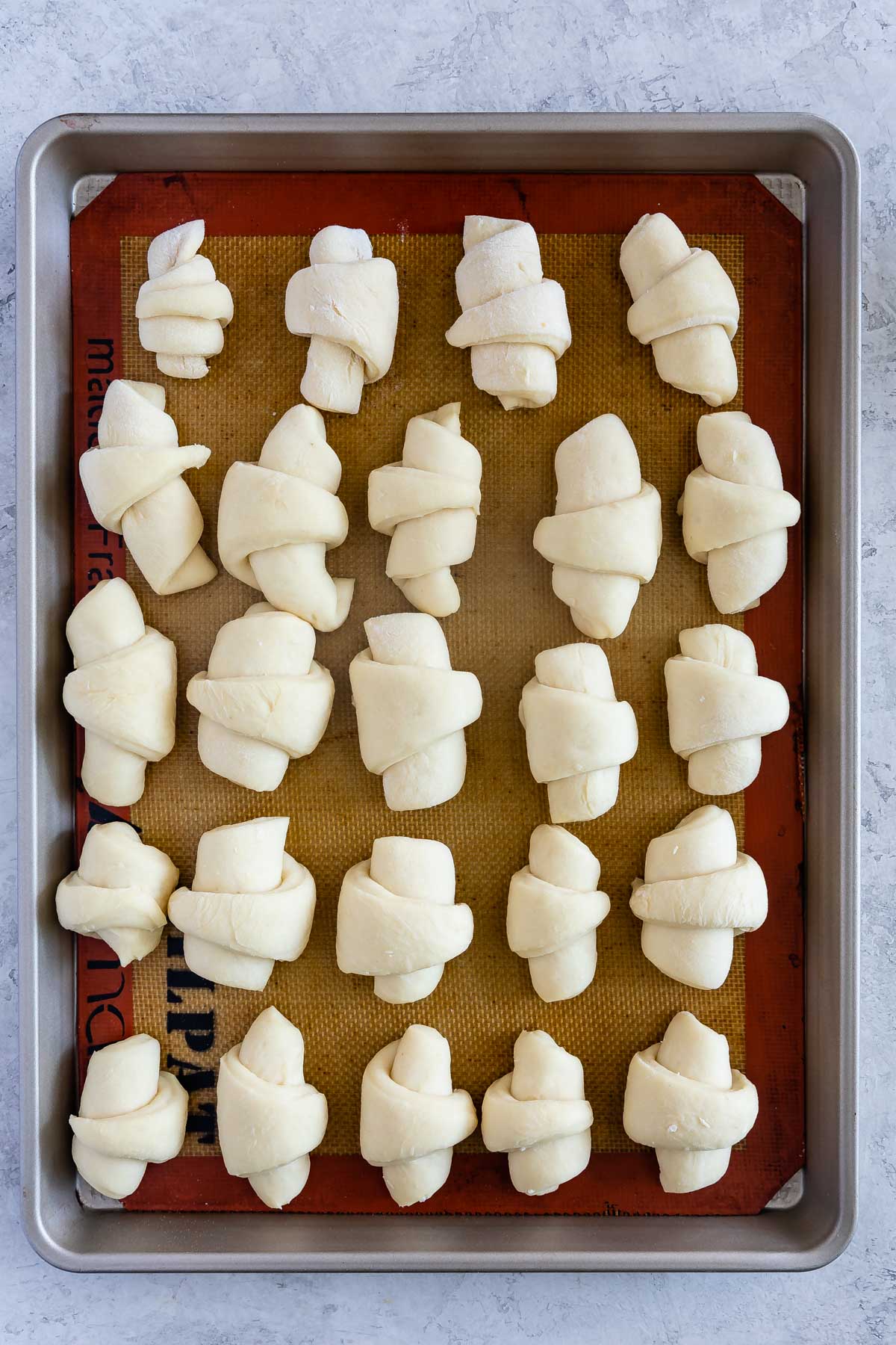 A baking sheet with 24 unbaked crescent rolls arranged in rows on a silicone baking mat. The rolls are evenly spaced and the dough is light in color.