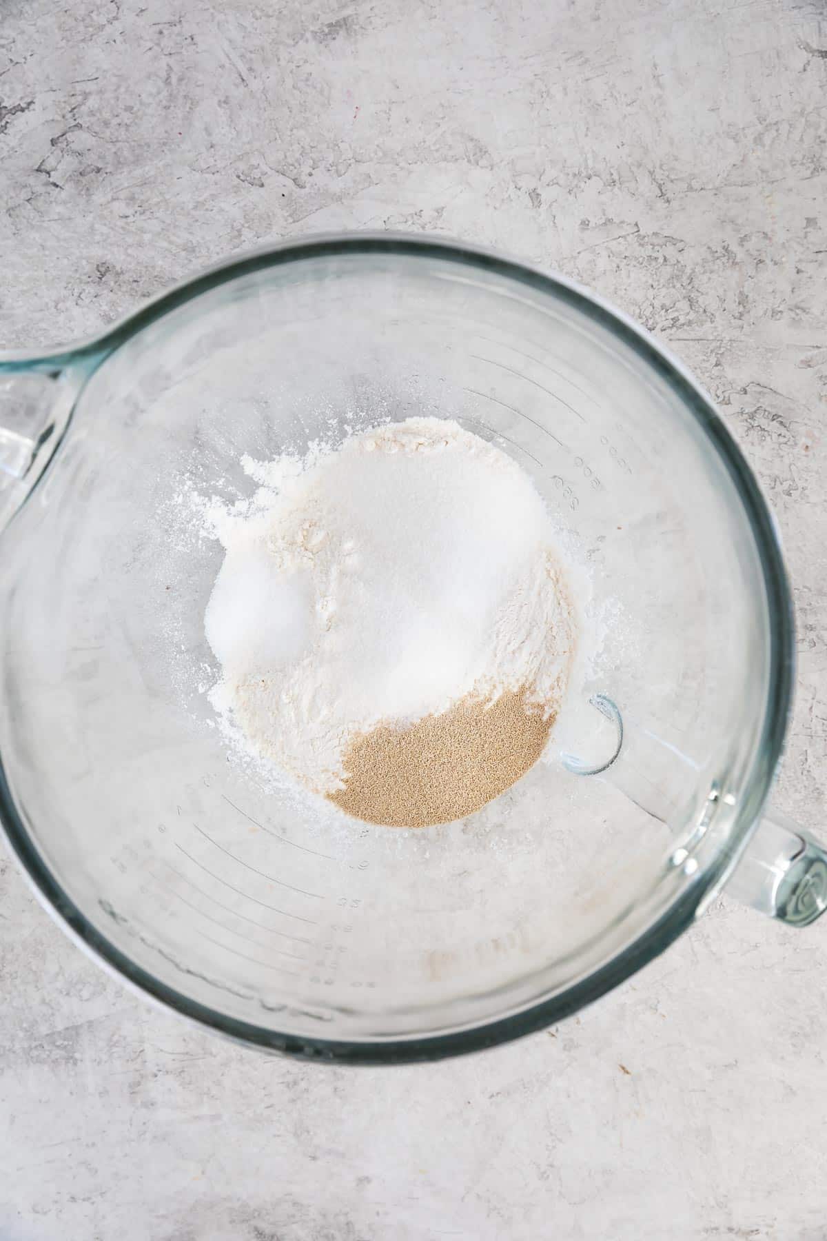 A glass mixing bowl containing flour, sugar, salt, and dry yeast on a light gray textured surface.