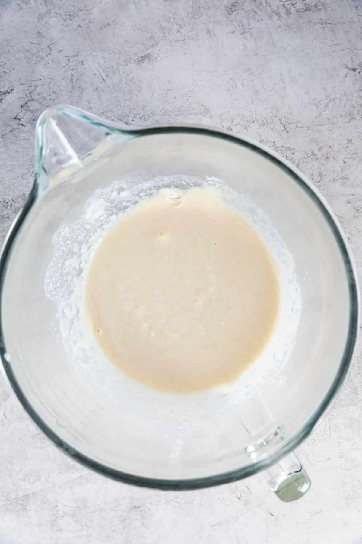 A glass mixing bowl with a pale, slightly frothy batter or liquid mixture sits on a light gray textured surface.