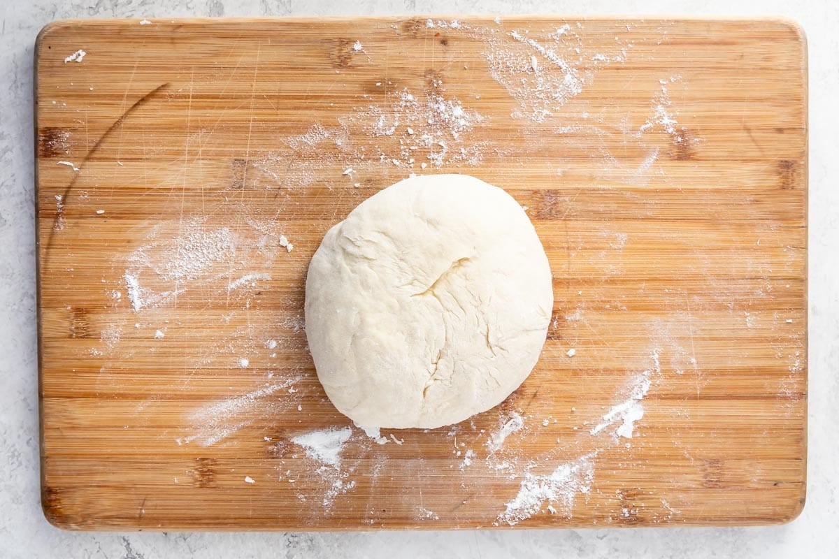A ball of dough sits on a wooden cutting board lightly dusted with flour, on a light-colored countertop.