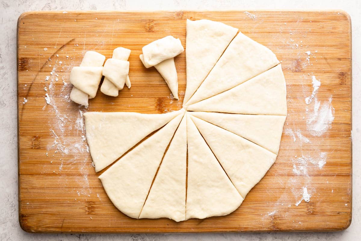 A wooden board holds dough cut into triangles, with three pieces rolled up and several unrolled pieces arranged in a circle. Flour is lightly scattered on the board.