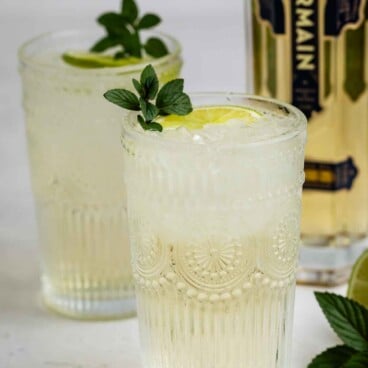 Two ornate glasses filled with a light-colored cocktail, garnished with mint and lime slices. A bottle of St-Germain liqueur and fresh mint leaves are visible in the background.