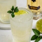 A glass of a pale cocktail with ice, garnished with a lime slice and mint sprig, sits on a white surface. A second glass, a bottle, and a lime half are in the background.