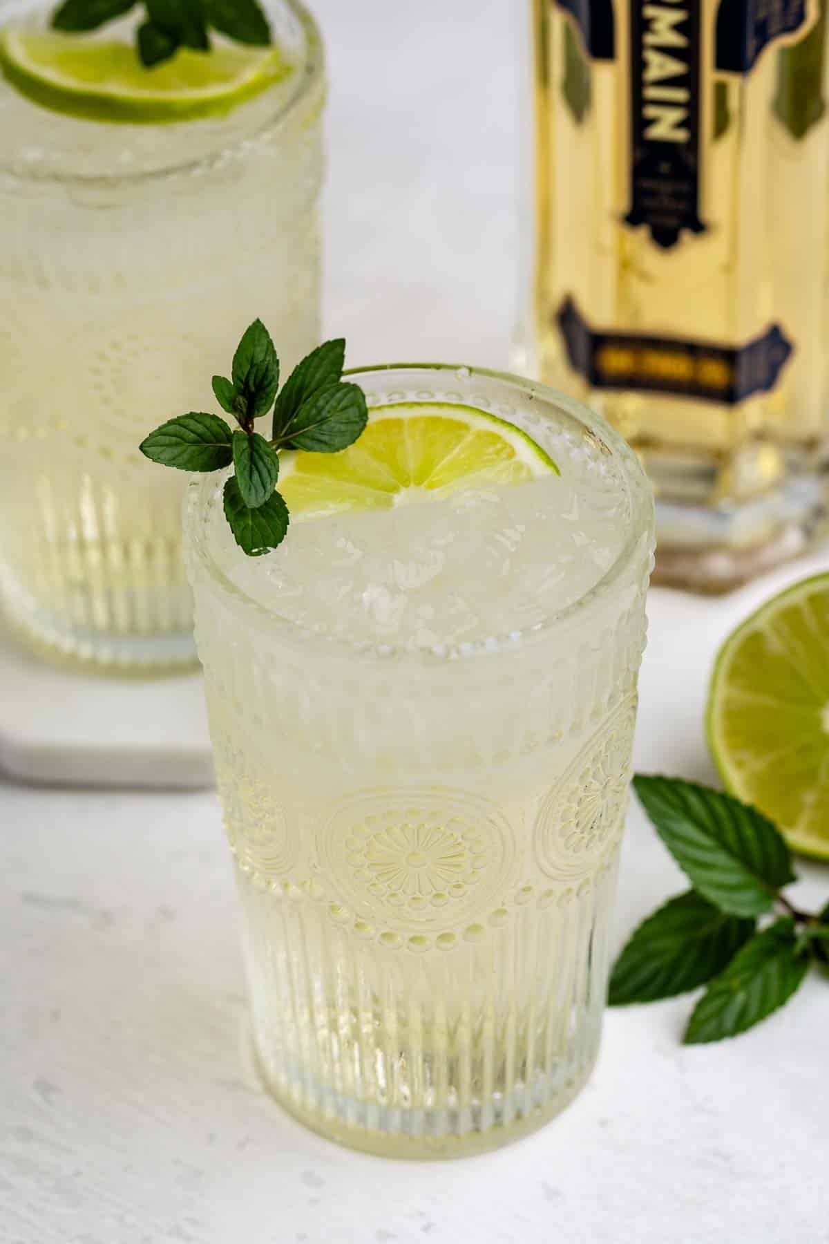 A glass of a pale cocktail with ice, garnished with a lime slice and mint sprig, sits on a white surface. A second glass, a bottle, and a lime half are in the background.