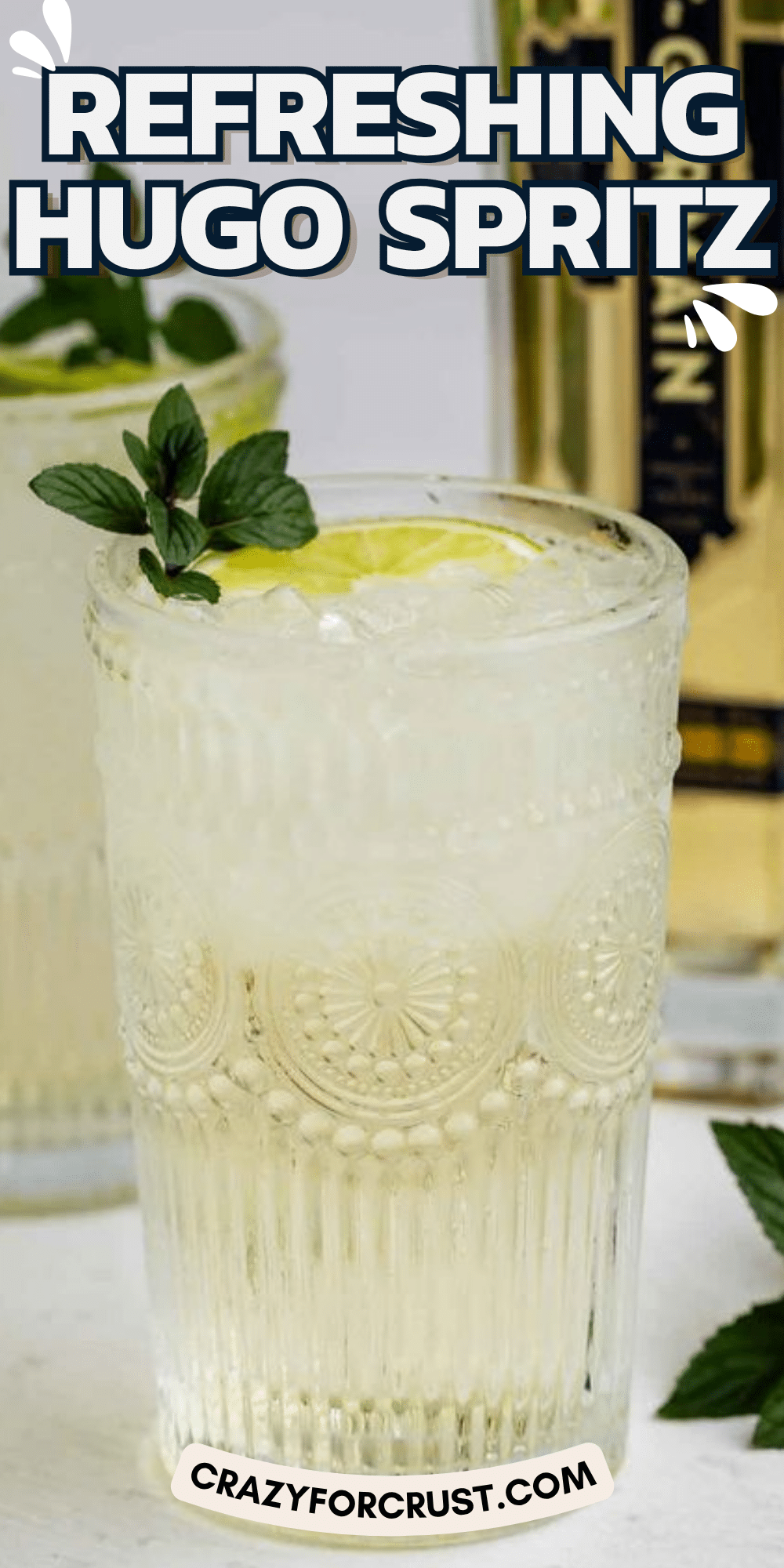 A glass of Hugo Spritz with ice, lime slice, and mint garnish sits on a white surface. Another glass and a bottle are blurred in the background. Text reads “Refreshing Hugo Spritz” and “crazyforcrust.com.”.