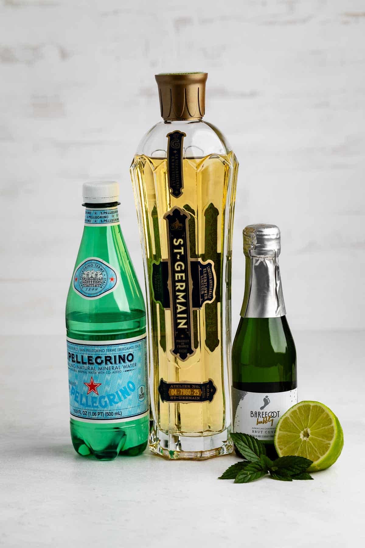 A bottle of St-Germain, a small bottle of Barefoot sparkling wine, a bottle of San Pellegrino, a halved lime, and fresh mint leaves are arranged on a white surface.