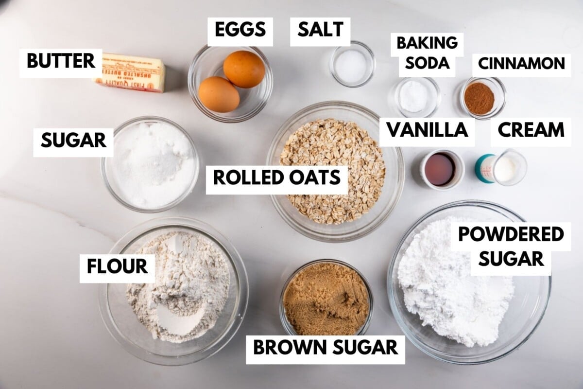 Top view of labeled baking ingredients in bowls on a white surface, including butter, sugar, flour, eggs, salt, rolled oats, brown sugar, powdered sugar, baking soda, cinnamon, vanilla, and cream.