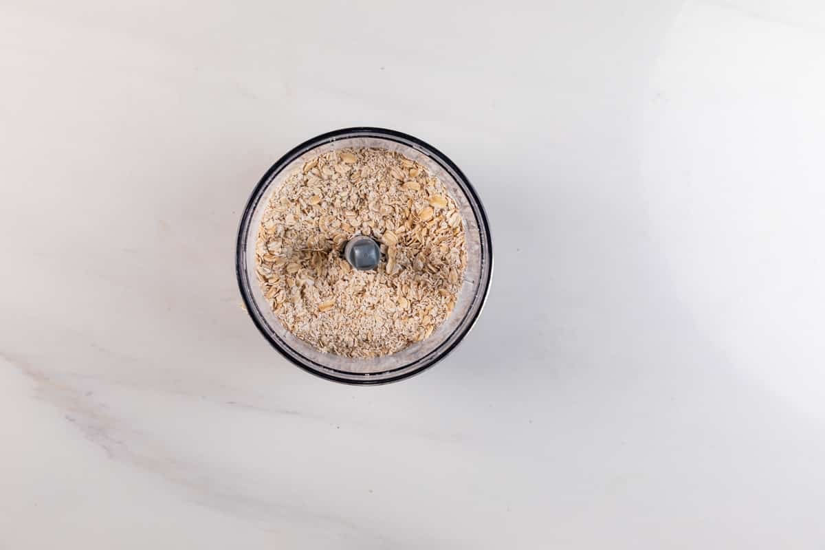 A top-down view of a food processor bowl filled with oats, placed on a white marble surface.
