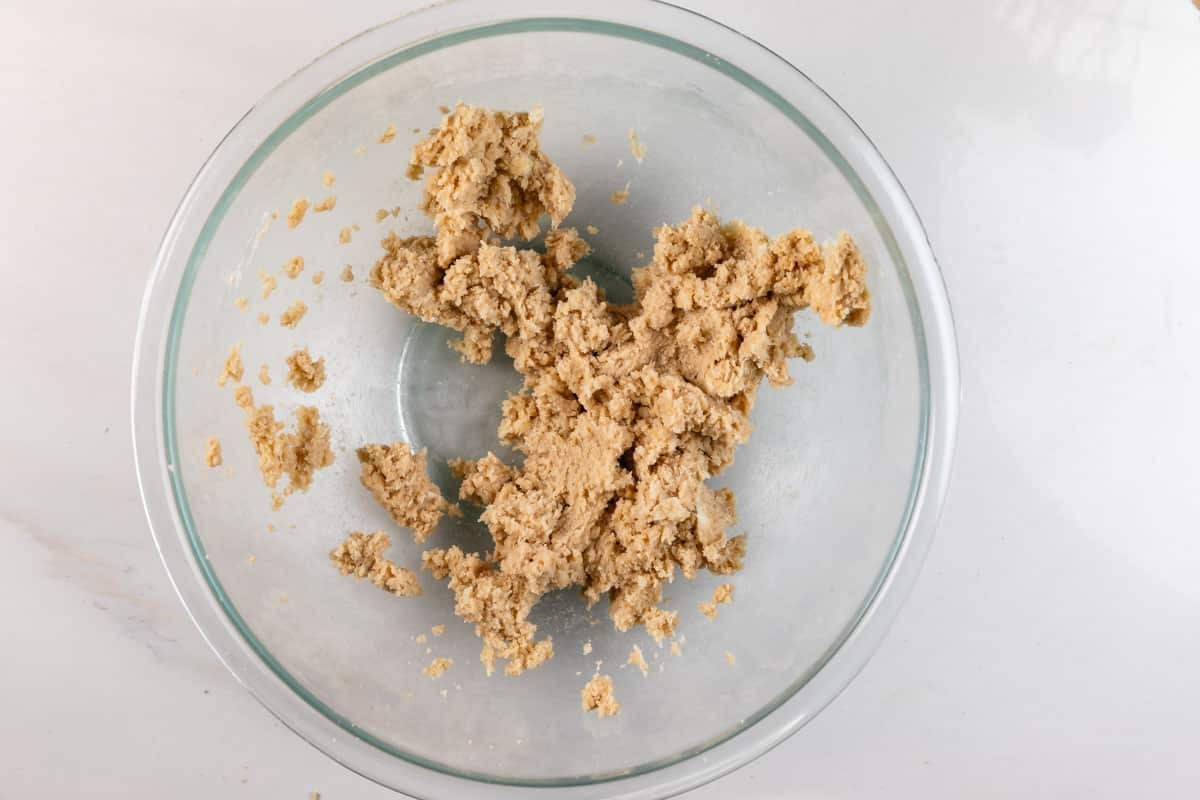 A glass bowl containing rough, beige cookie dough sits on a white surface. Some dough is clumped together in the center, with small bits scattered around the edges of the bowl.