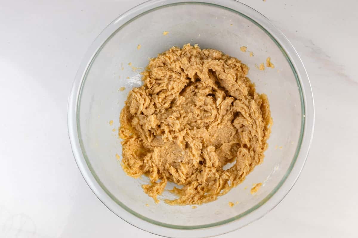 A glass bowl containing thick, light brown cookie dough sits on a white surface.