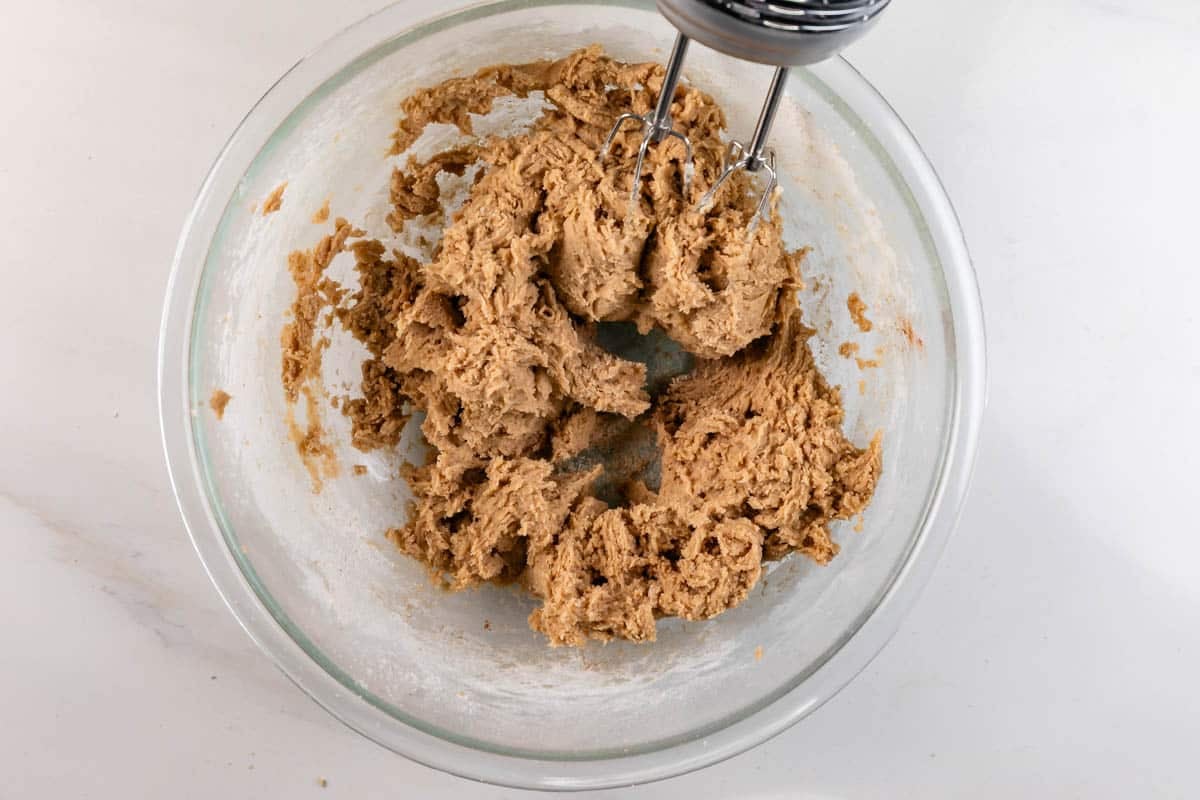 A glass bowl with thick cookie dough being mixed by an electric hand mixer on a white surface.