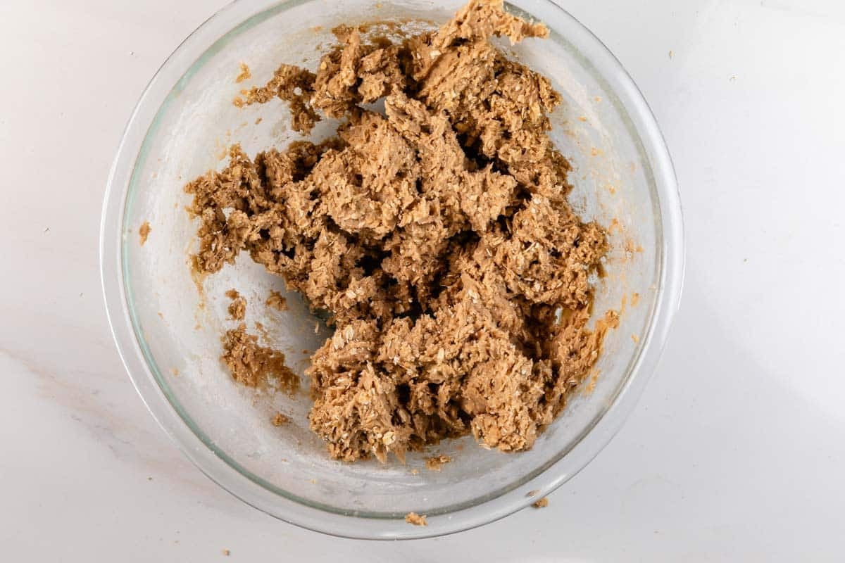 A glass bowl filled with chunky, brown cookie dough sits on a white surface.