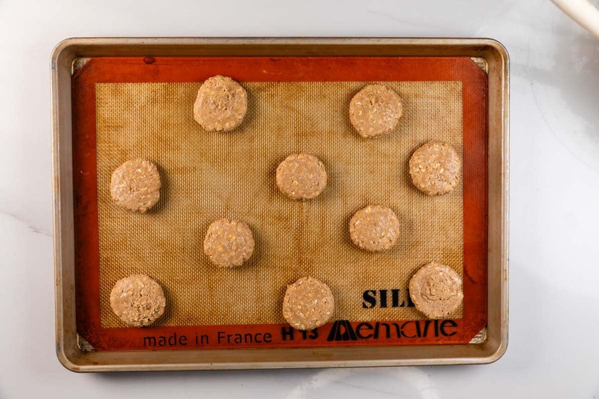 A baking sheet with a silicone mat holds ten evenly spaced, unbaked oatmeal cookie dough balls ready to be baked.
