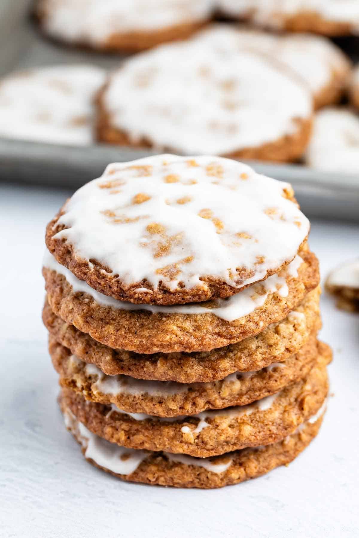 A stack of six oatmeal cookies with white icing on top, with more cookies blurred in the background on a tray.