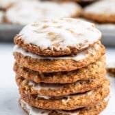 A stack of five oatmeal cookies with white icing on top, sitting on a white surface, with more cookies blurred in the background.