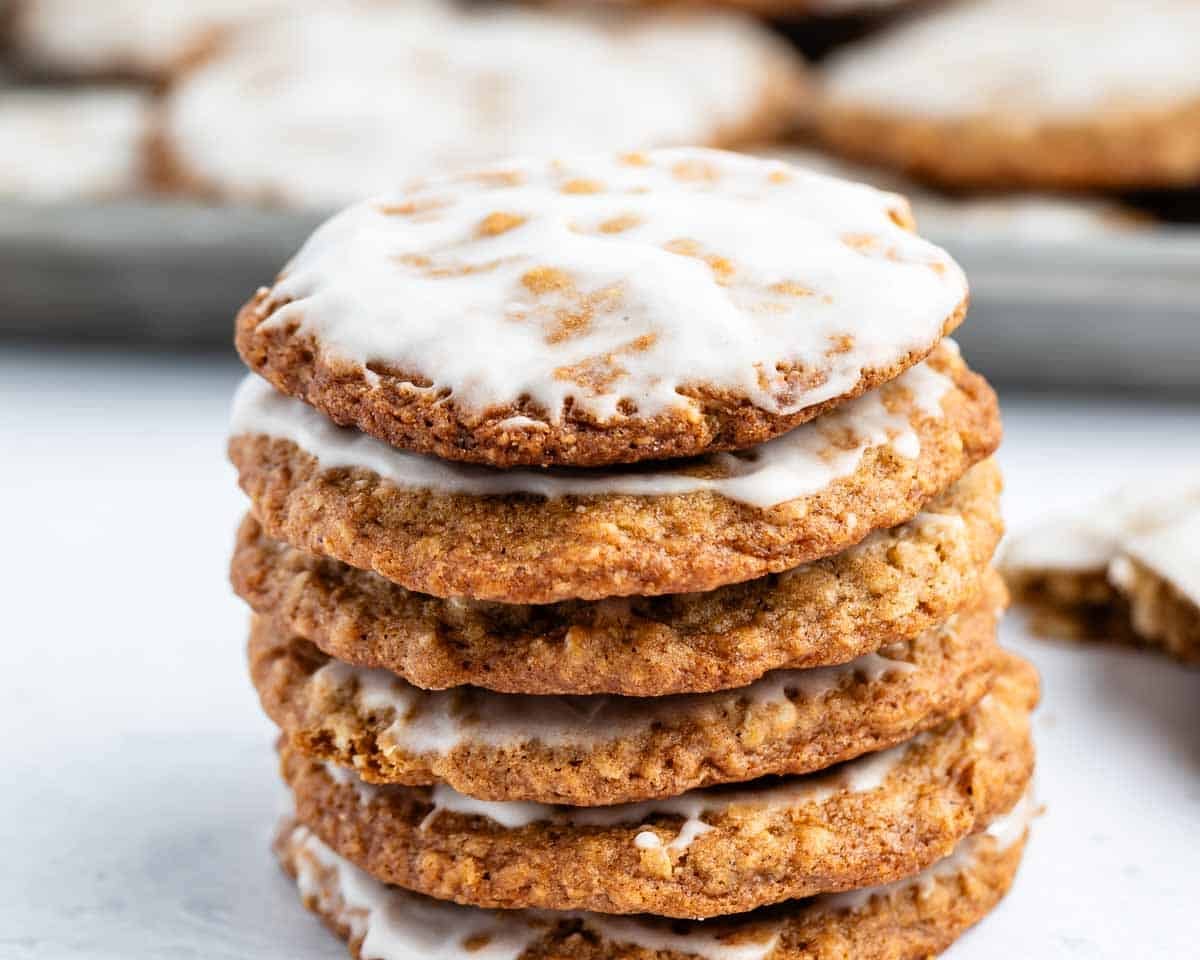 A stack of five oatmeal cookies with white icing on top, sitting on a white surface, with more cookies blurred in the background.